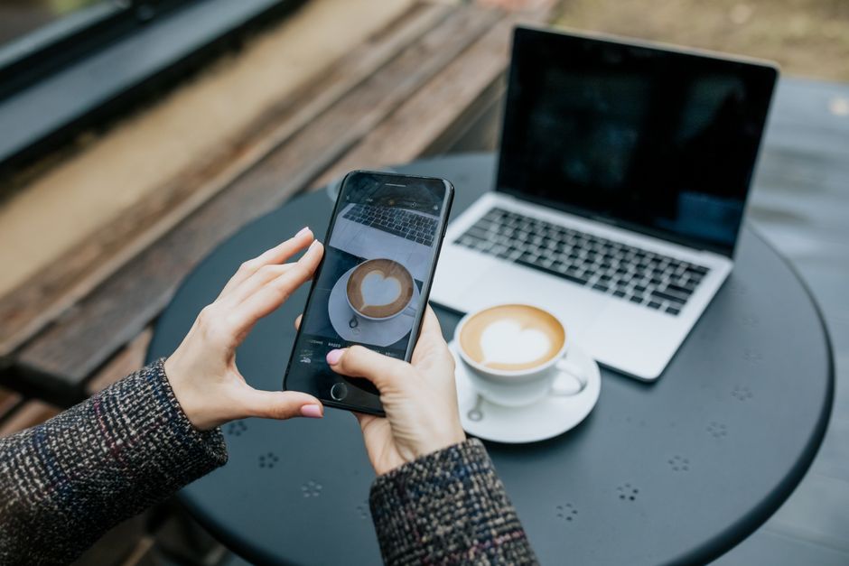 カフェのテーブルでスマートフォンを構え、ラテの写真を撮っている様子のイメージ画像