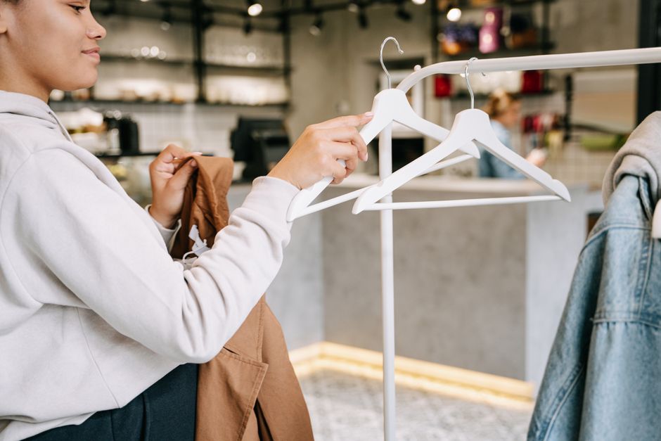 ハンガーラックに服が並ぶ小さな服屋の店内で、客が1着を手に取って選んでいるイメージ画像
